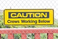 Closeup of a yellow [Caution Crews Working Below] sign on a chain-link fence under the sunlight