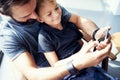 Closeup of young boy sitting with father and using mobile phone in modern sunny place. Horizontal, blurred background.