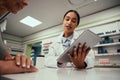 Young female pharmacist hand holding digital tablet wearing labcoat and standing behind counter guiding customer