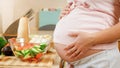 CLoseup of young pregnant woman cooking and eating vegetable salad holding big abdomen and touching it with hands Royalty Free Stock Photo