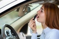 Closeup of a young redhead woman driver correcting her makeup with dark red lipstick looking in car rearview mirror behind Royalty Free Stock Photo