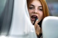 Closeup of a young redhead woman driver correcting her makeup with dark red lipstick looking in car rearview mirror behind Royalty Free Stock Photo