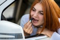 Closeup of a young redhead woman driver correcting her makeup with dark red lipstick looking in car rearview mirror behind Royalty Free Stock Photo
