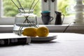 A clothed table has a vase, some lemons on a plate and a magazine placed in the natural light of the windows as part of a home