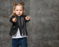 Little cute blond girl in stylish rock style black leather jacket, jeans and sneakers standing and pointing at camera with fingers Royalty Free Stock Photo