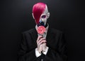 Clown and Halloween theme: Scary clown with pink hair in a black jacket with candy in hand on a dark background in the studio Royalty Free Stock Photo