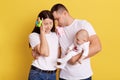 Clueless mother and father with baby posing isolated over yellow background, couple keep eyes closed, dad leans on wife`s head,