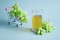 Clusters of fresh white grapes next to a miniature supermarket cart filled with grapes and a clear glass of grape juice. Blue
