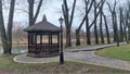 A cobblestone and wood-paved path approaches the wooden gazebo, and next to it is a metal pole with a lantern. Behind them grow ol