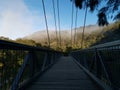 Beautiful early morning view of a suspension bridge with blue sky, light clouds, mountains, trees and fog in the background Royalty Free Stock Photo