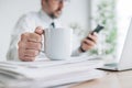 Coffee break at workplace, businessman drinking coffee and using smartphone in office