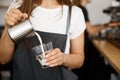 Coffee Business Concept - close-up lady barista in apron preparing and pouring milk in glass cup while standing at cafe. Royalty Free Stock Photo