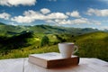A coffee cup and a book on a wooden table against the backdrop of a beautiful green mountain landscape. Royalty Free Stock Photo