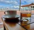 Coffee Cup and glass of Whisky (Whiskey) on table with blue cloudy sky behind. Royalty Free Stock Photo