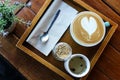 Coffee cup with heart shape latte art in wooden tray with brown sugar, spoon, tea, napkin, and decorating flower Royalty Free Stock Photo