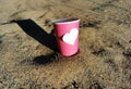 Coffee cup standing in the send near the road. Pink paper cup with blank heart shape to add some text. Joyful template for romanti