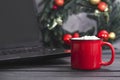 Coffee or hot chocolate and marshmallows, next to a spruce Christmas wreath with red and gold balls and a laptop on a gray Royalty Free Stock Photo