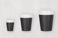 Coffee mockup - set of three different size black paper cups and blank white caps on white wood board, top view. Royalty Free Stock Photo