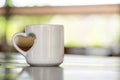 A coffee mug with a love shaped handle standing on the top of a table with blurred out bright background