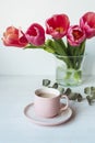 Coffee in pink cup and bouquet of tulip flowers in glass vase on white table. Spring holidays concept. Close up Royalty Free Stock Photo