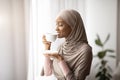 Coffee time. Gorgeous black woman in hijab sipping hot aromatic drink near window at home Royalty Free Stock Photo