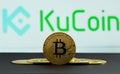 The coins of bitcoin are in front of logo of Ku Coin crypto stock exchange background. In the foreground is a BTC coins and on the