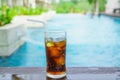 Cola with ice in glass and white straw on wood near swimming pool