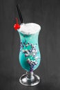 Cold alcoholic blue cocktail, decorated with berries and flowers in glass on black wooden background