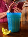 Cold beverage in blue plastic glass with lemon wedge and hands using phone Royalty Free Stock Photo