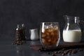 Cold coffee in a glass with ice cubes, where milk is subsequently poured on dark gray background