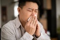 Cold, flu, allergy concept. Sick middle aged asian man sneezing in napkin, ill male feeling unwell, sitting on couch Royalty Free Stock Photo