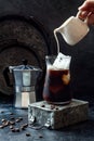 Cold refreshing iced coffee in a tall glass and coffee beans on dark background. Pouring cream into glass with iced coffee. Royalty Free Stock Photo