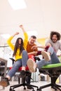 Colleagues having fun on work at the office. Girls pushing boys on chairs with wheels. Multiethnic group of successful start up