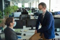 Colleagues in medical masks in an open office space communicate at the desk. A man and a woman in office suits are