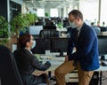 Colleagues in medical masks in an open office space communicate at the desk. A man and a woman in office suits are