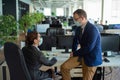 Colleagues work in medical masks in an open office space communicate at the desk. A man and a woman in office suits are