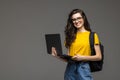 College student girl on gray background, smiling at camera, pressing laptop to chest, wearing backpack, ready to go to studies, Royalty Free Stock Photo