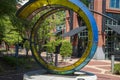 A colorful circular glass sculpture at the Waterhouse Pavilion with red brick buildings and sidewalks, lush green trees and plants