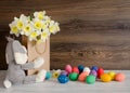 Colorful Easter eggs with lovely bouquet of narcissus in paper bag and funny bunny on wood background