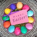 Colorful Easter Eggs on a Plate in center with Pink Happy Easter Name Tag Card on Vintage Lace Tablecloth. A Square crop with goo