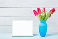 Colorful red spring tulip flowers in nice blue vase and blank photo frame on light wooden background as greeting card. Mothersday