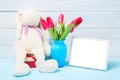 Colorful red spring tulip flowers in nice blue vase, blank photo frame and stuffed toy bunny on light wooden background as greetin Royalty Free Stock Photo