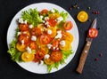 Colorful salad, fresh green leaves and sliced red and yellow cherry tomatoes, white plate, knife, black stone background, top view Royalty Free Stock Photo