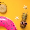 Colorful flat lay with pink inflatable circle donut, funny pineapple in sunglasses, bamboo bag and starfish over yellow background