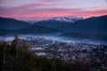 Colorful sunset over snow covered mountains and city with fog. Trees in foreground. Royalty Free Stock Photo