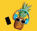 Colorful trendy image of stylish pineapple in wireless headphones listening to music with phone over yellow background