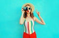 Colorful young woman holding retro camera, blowing red lips sends air kiss in summer straw hat having fun on blue wall