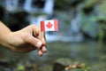 Colour man has in hand national flag of Canada on wooden stick. He waves with state symbol near a river. In the background are