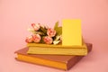 Colourful books, flowers and blank yellow paper card for message on pink background. Spring home decor.