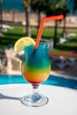 Colourful cold Rainbow Paradise cocktail drink served in glass at pool bar overlooking blue pool, sea and palm trees, relax and